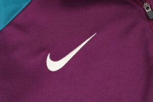 White Nike swoosh logo on purple fabric (likely backpack or jacket) with a teal corner and zipper edge behind it.
