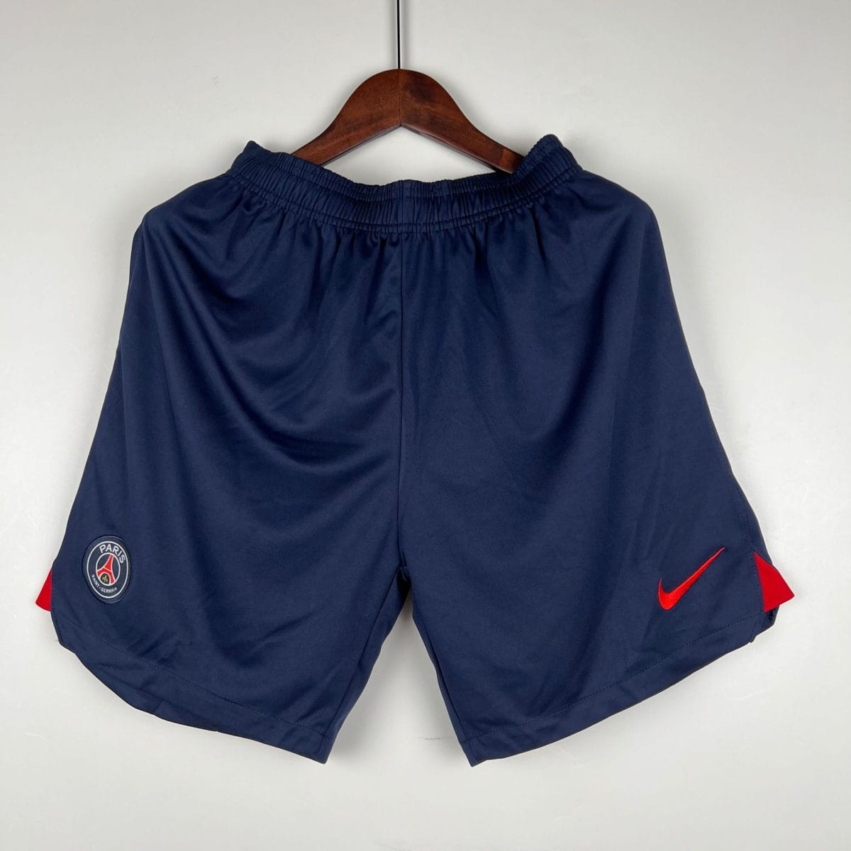 Navy athletic shorts on a hanger with PSG badge on the left leg and a red Nike swoosh on the right leg.