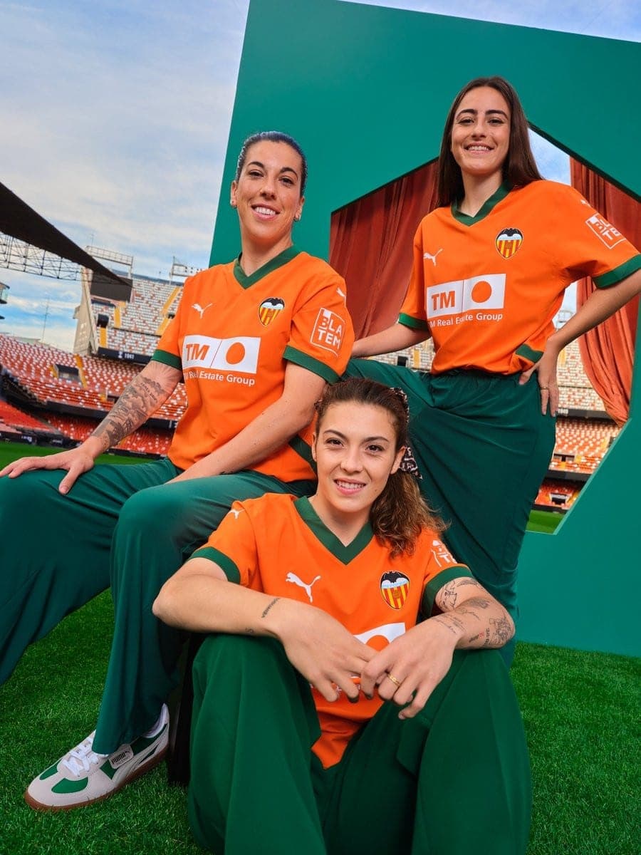 80kJUgdbBIpv71P | UAE SPORT STORE Three Valencia CF players in orange jerseys and green pants posing together on a football pitch with stadium seating behind them.