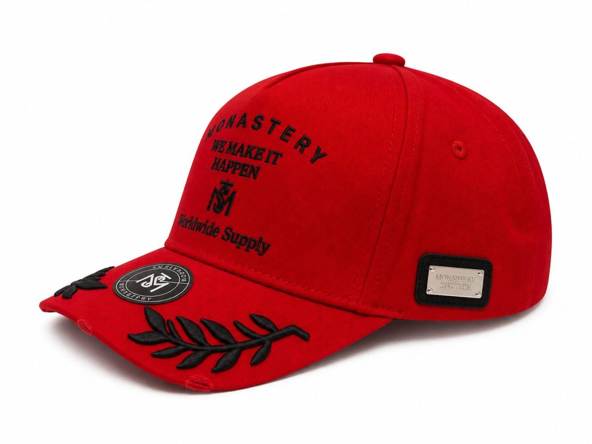 Red cap with black embroidery saying 'We Make It Happen' and decorative black leaf motif on the brim; metal plaque on the side.
