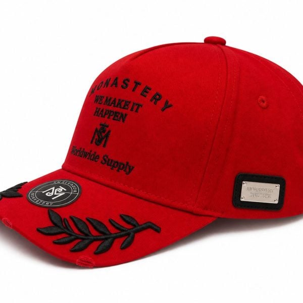 Red cap with black embroidery saying 'We Make It Happen' and decorative black leaf motif on the brim; metal plaque on the side.