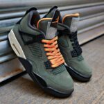 Pair of olive-green sneakers with bright orange laces resting on a metal surface in an urban setting.