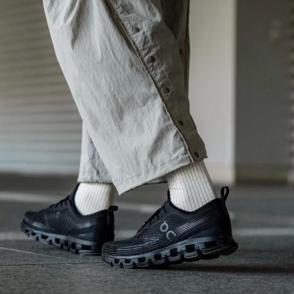 Person wearing black athletic sneakers with chunky soles, white ribbed socks, and beige wide-leg pants walking indoors on a concrete floor.