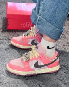Pair of pink Nike sneakers with white laces and rhinestone embellishments, worn with rolled-up jeans and white Nike socks on a gray carpeted floor, pink Nike box in the background