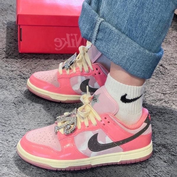 Pair of pink Nike sneakers with white laces and rhinestone embellishments, worn with rolled-up jeans and white Nike socks on a gray carpeted floor, pink Nike box in the background