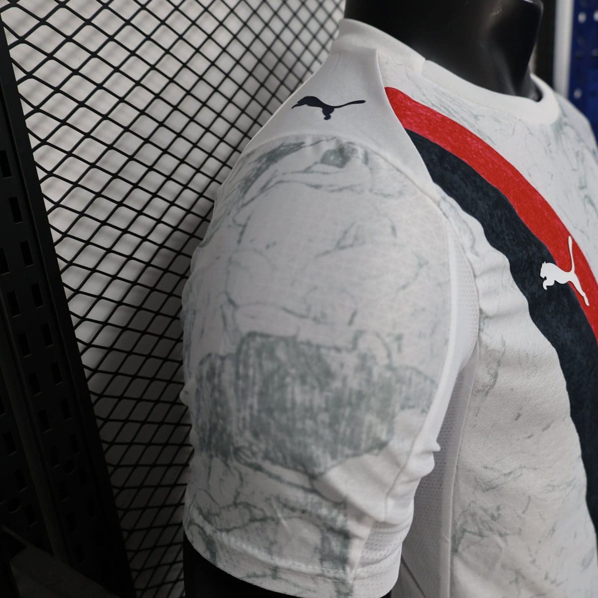 Close-up of a white sports jersey with a red and navy graphic and a small Puma logo on the shoulder, against a metal grid backdrop.