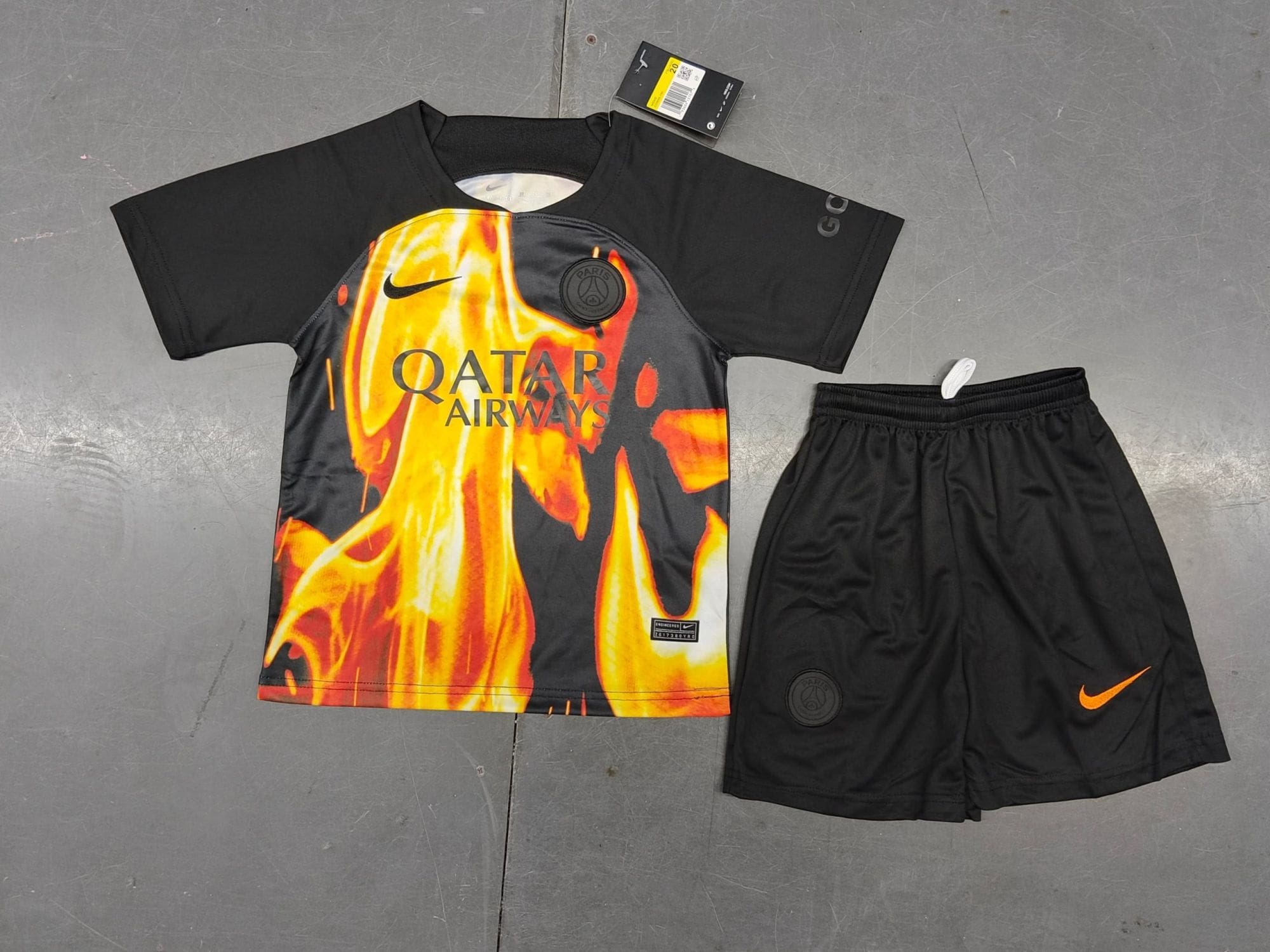 82452201_a97dd951-4d07-4920-92e9-e77f02d10dcd | UAE SPORT STORE Black Paris Saint‑Germain football jersey with a fiery orange flame design and Qatar Airways branding, paired with black shorts with an orange Nike swoosh on a gray floor.