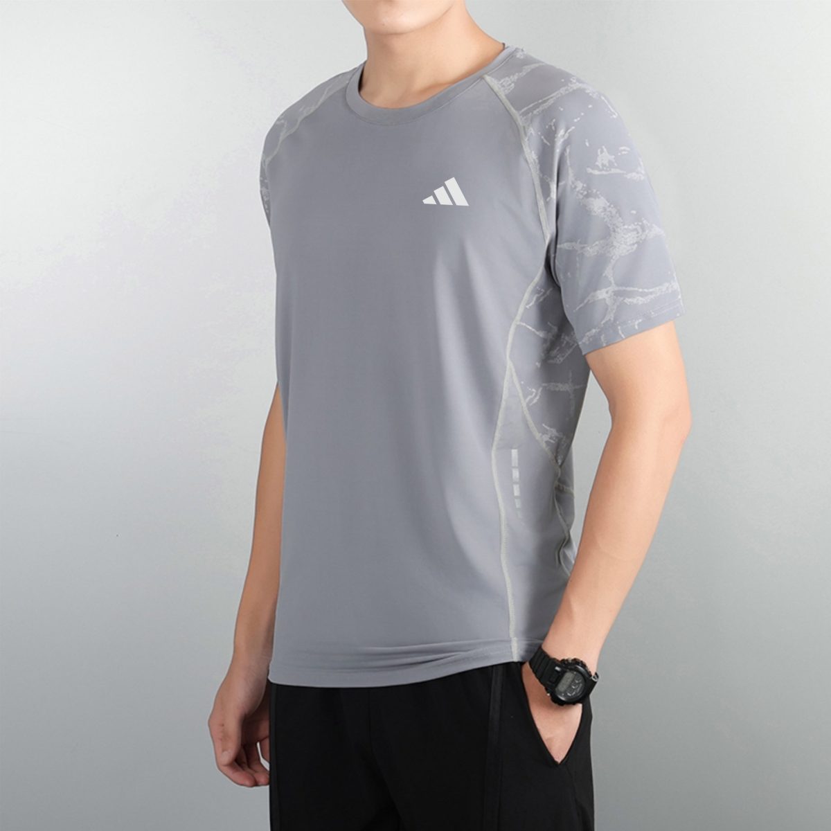 Person in a gray athletic shirt with a small white logo on the chest, standing against a pale wall, wearing a black watch on the left wrist.