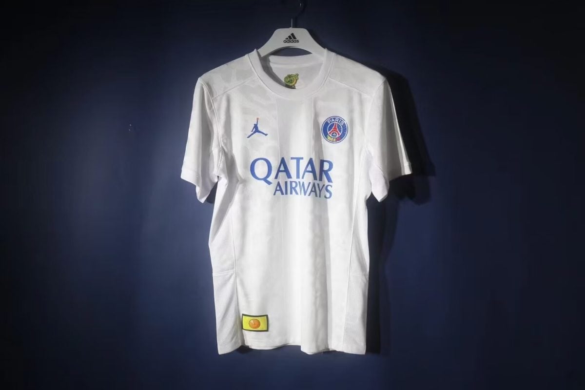 White PSG football jersey on a hanger with Qatar Airways sponsor and blue club crest, displayed against a dark background.