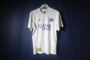 White PSG football jersey on a hanger with Qatar Airways sponsor and blue club crest, displayed against a dark background.
