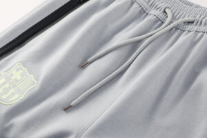 Close-up of light gray joggers showing drawstrings, elastic waistband, and a neon-green shield logo on the left leg/pocket area.