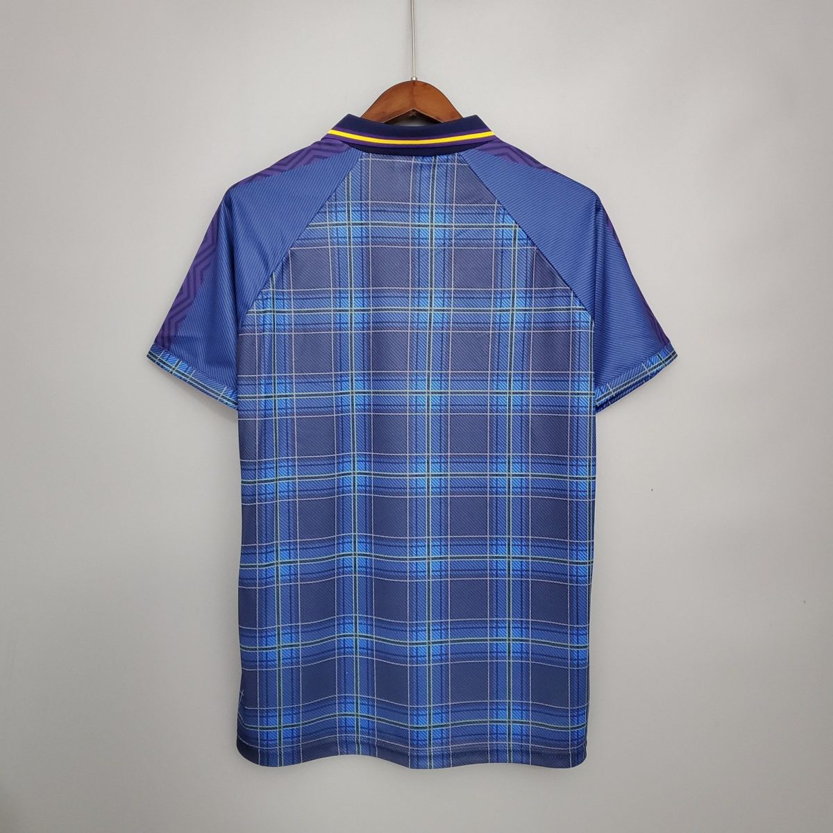 Blue plaid shirt or jacket on wooden hanger, back view, hanging against a light wall with purple sleeves and yellow trim on the collar.