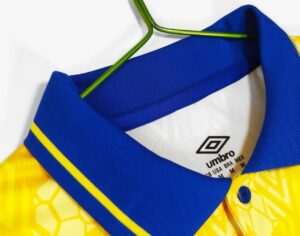Close-up of a blue polo shirt collar with yellow trim on a green hanger; Umbro label visible inside the neck area, yellow patterned shirt underneath.