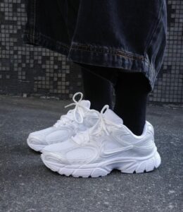 White chunky sneakers on a person’s feet, wearing dark jeans and black socks, standing on a concrete pavement.