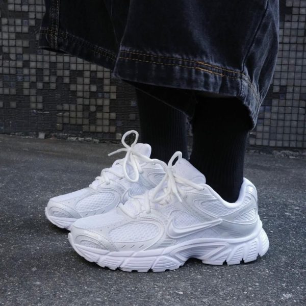 White chunky sneakers on a person’s feet, wearing dark jeans and black socks, standing on a concrete pavement.