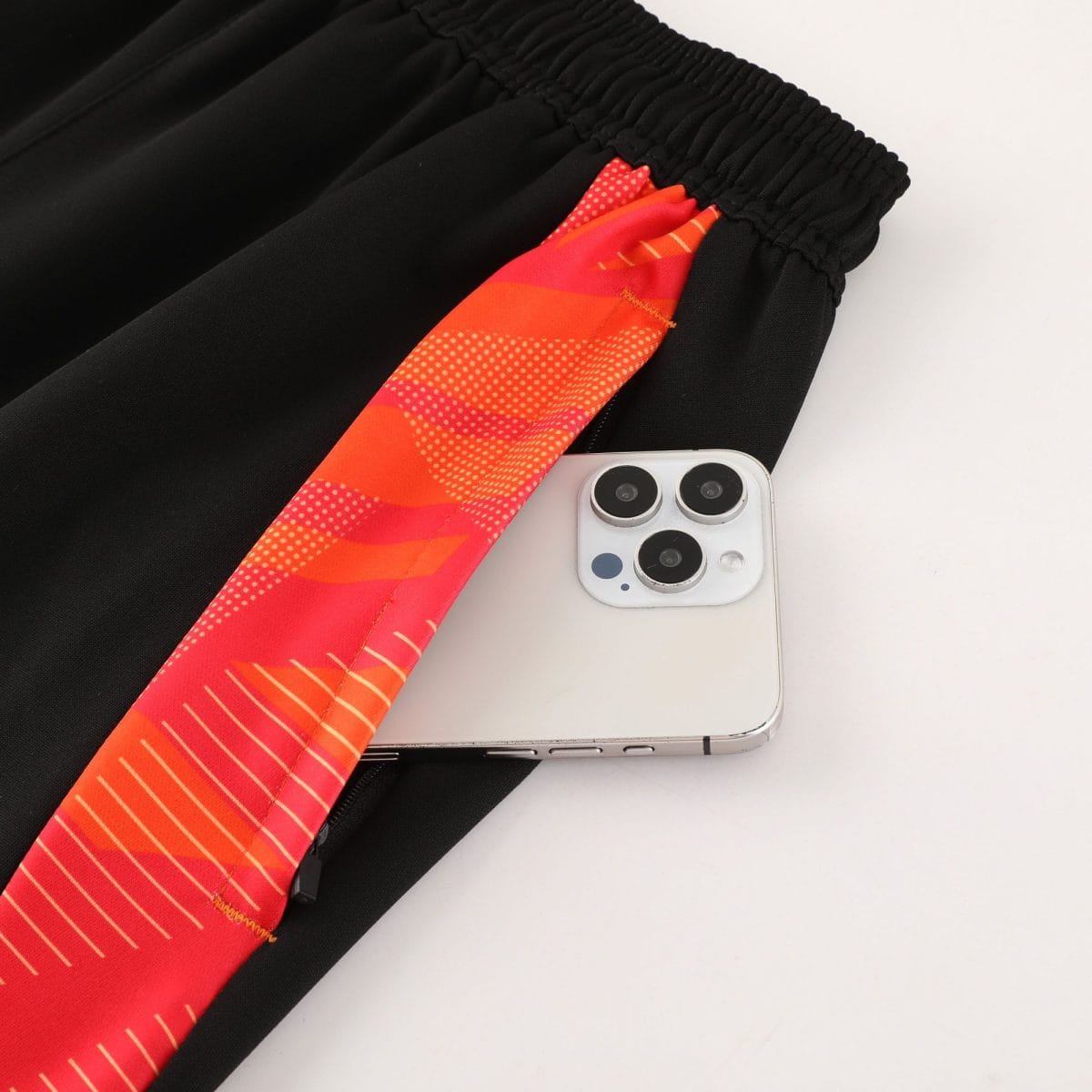 Close-up of a black fabric with an elastic cuff and a bright orange patterned strap lying across it, next to a silver smartphone with three cameras.