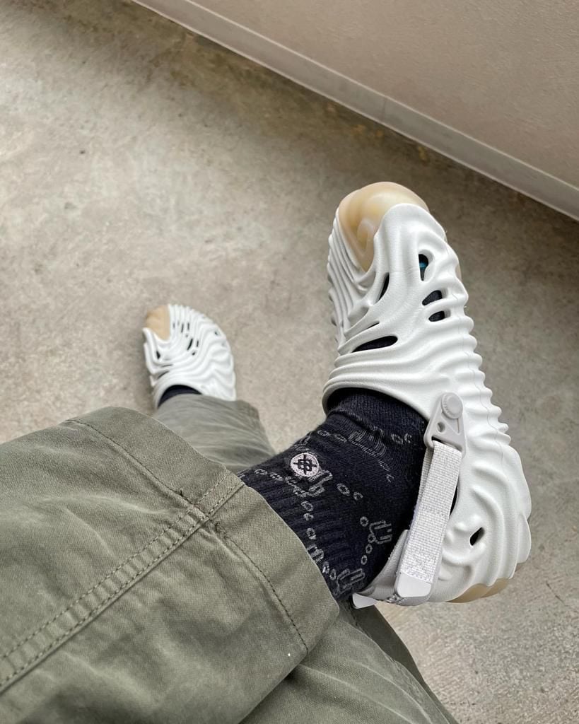 8339cf2a-cf88-4880-b170-9cc7cd96c9ea | UAE SPORT STORE Person wearing white sculpted sandals over black socks, olive-green pants, standing on a concrete floor.