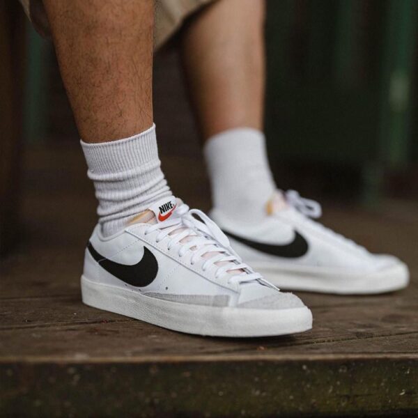 Person wearing white Nike sneakers and white socks standing on a wooden floor, close-up of the shoes' sides and logo