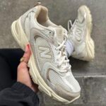 8472879a-648f-4d97-8bdf-f03def2a1a43 | UAE SPORT STORE Beige New Balance running shoe with mesh upper, held in hand; second shoe visible on pavement in background.
