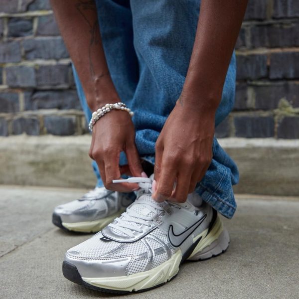 Person tying the laces of white athletic sneakers while wearing blue jeans and pearl bracelets, outdoors against a brick wall.