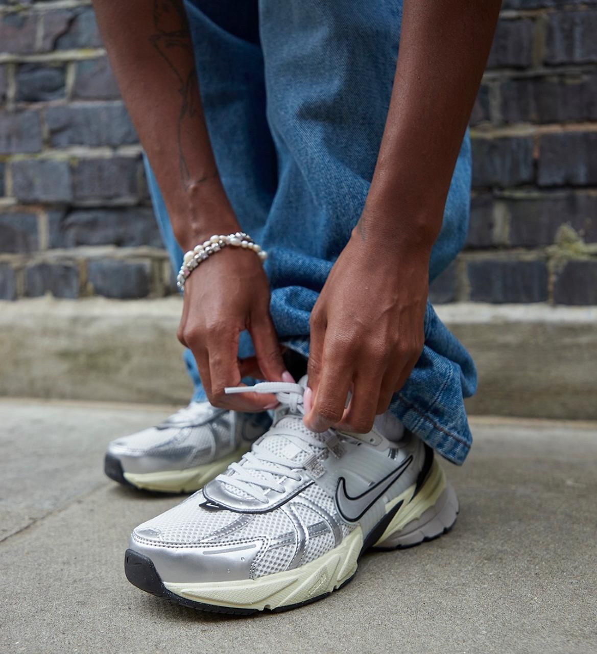 85928396-0384-46ed-b92e-0c04e3748785 | UAE SPORT STORE Person tying the laces of white athletic sneakers while wearing blue jeans and pearl bracelets, outdoors against a brick wall.