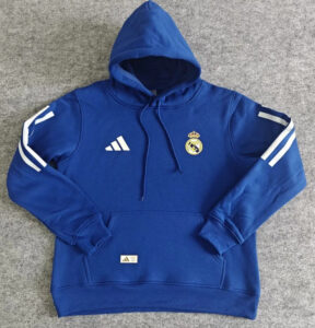 Blue Real Madrid hoodie with hood, features club crest and Adidas logo, white sleeve stripes, and a front pocket on a gray background.