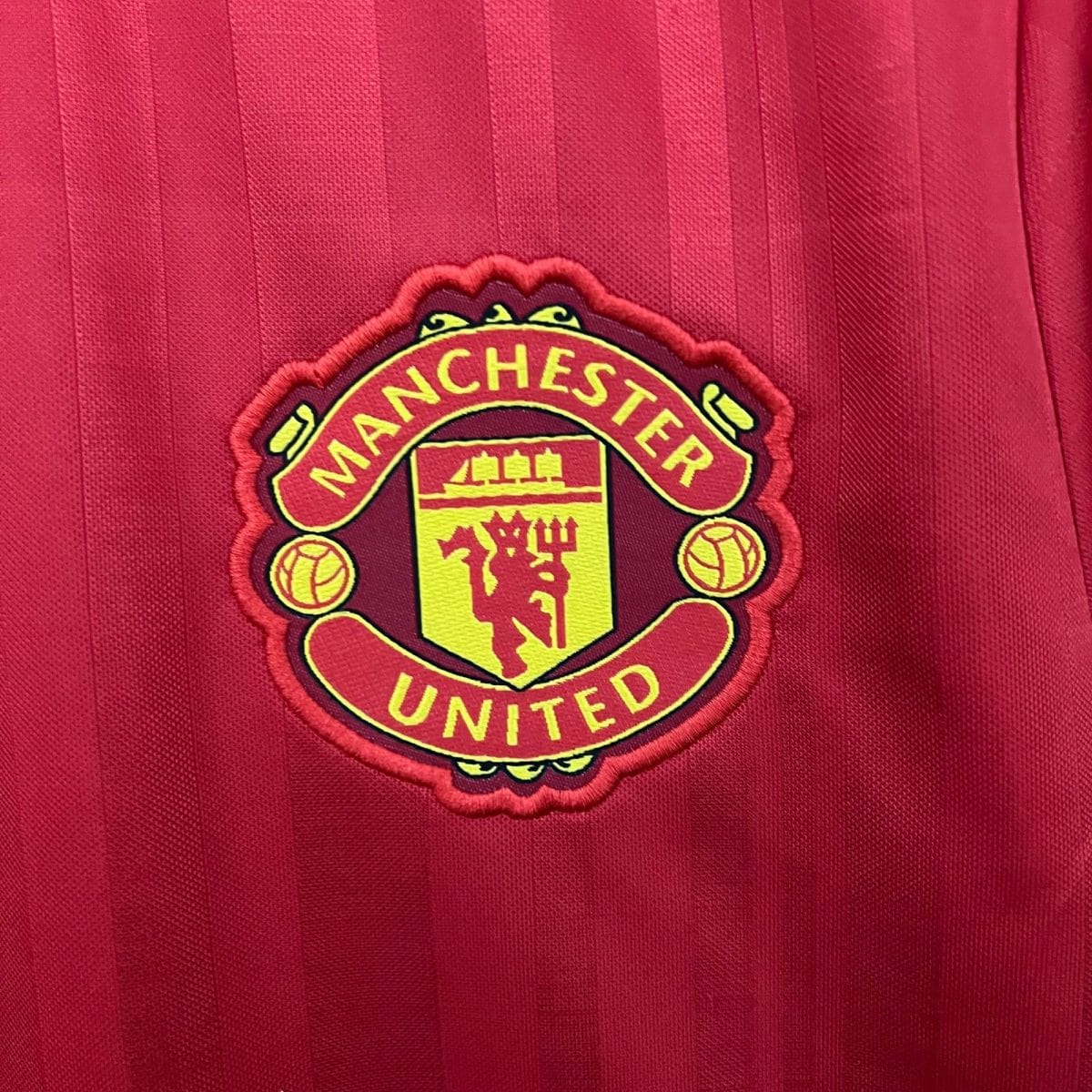 Manchester United crest on a red striped jersey fabric, close-up detail of the badge edge on the left side of the image