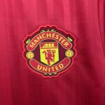 85f9ff0d_5a54c02c-60df-4f4e-b730-5eb11978a341 | UAE SPORT STORE Manchester United crest on a red striped jersey fabric, close-up detail of the badge edge on the left side of the image