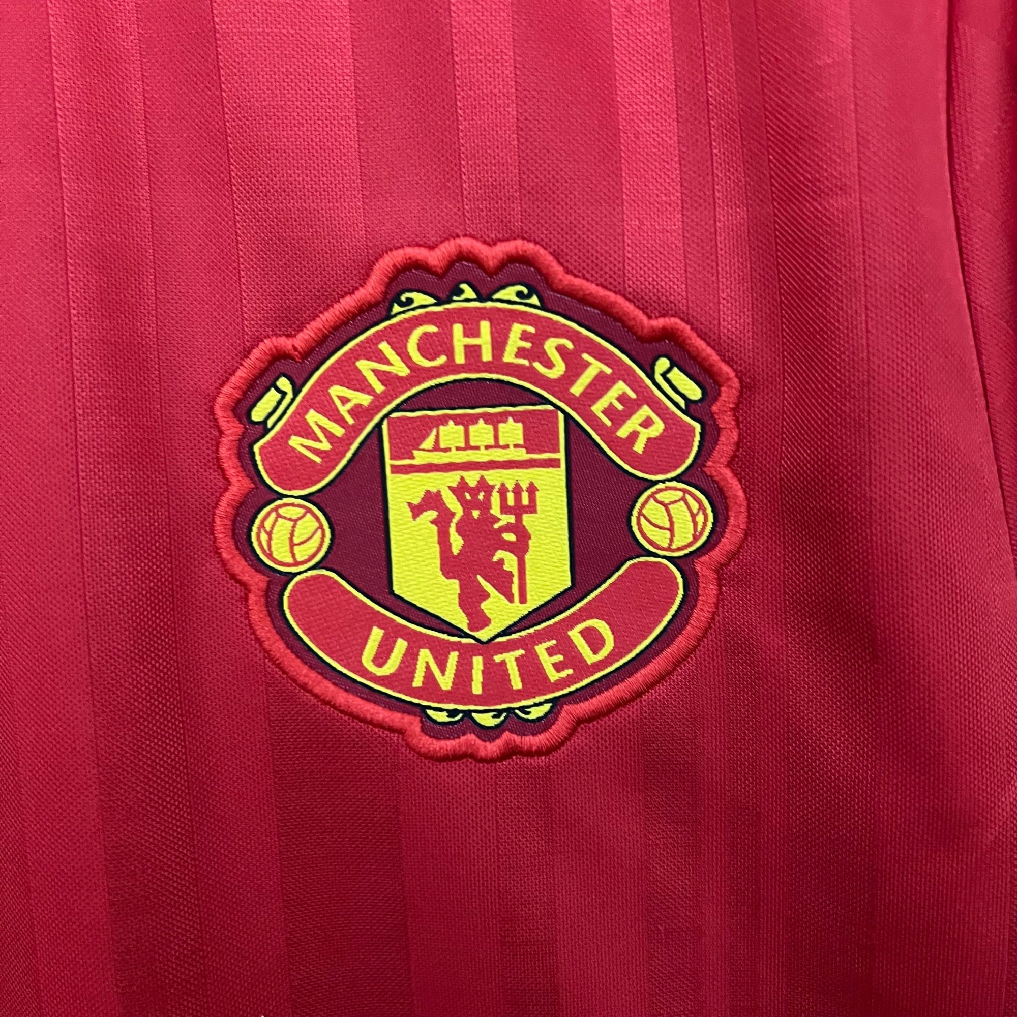 85f9ff0d_5a54c02c-60df-4f4e-b730-5eb11978a341 | UAE SPORT STORE Manchester United crest on a red striped jersey fabric, close-up detail of the badge edge on the left side of the image