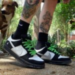 Person wearing black-and-white sneakers with bright green laces and black socks, standing outdoors with a dog in the background.