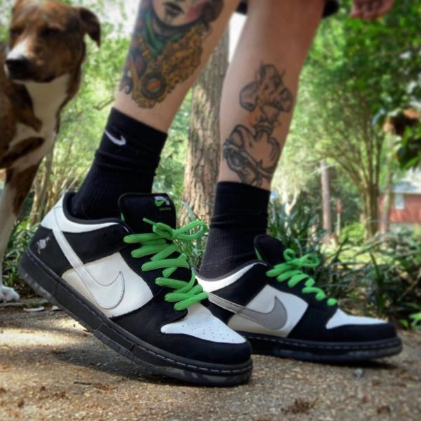Person wearing black-and-white sneakers with bright green laces and black socks, standing outdoors with a dog in the background.
