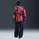 Person facing away, wearing a neon red and black star-pattern jacket with 'STATES' on the back, black pants, and a cap in a studio.
