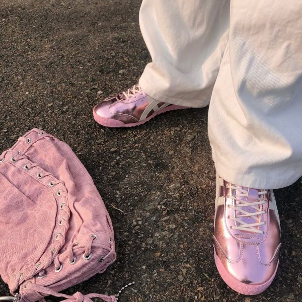 A person wearing pink metallic sneakers and white pants stands on asphalt next to a pink lace-up bag on the ground.