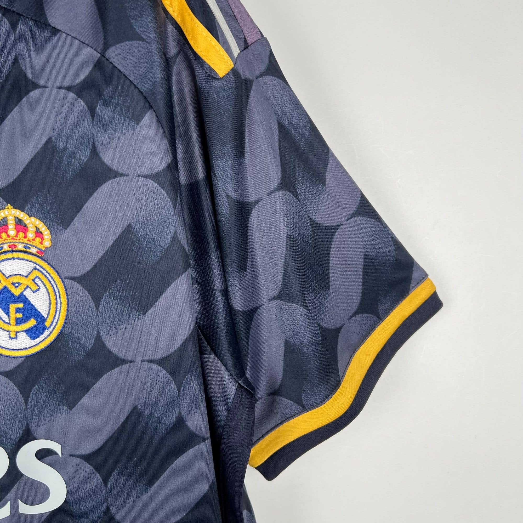 872c8303 | UAE SPORT STORE Close-up of a dark blue patterned athletic jersey with yellow trim and part of a circular team crest visible at the bottom left corner.