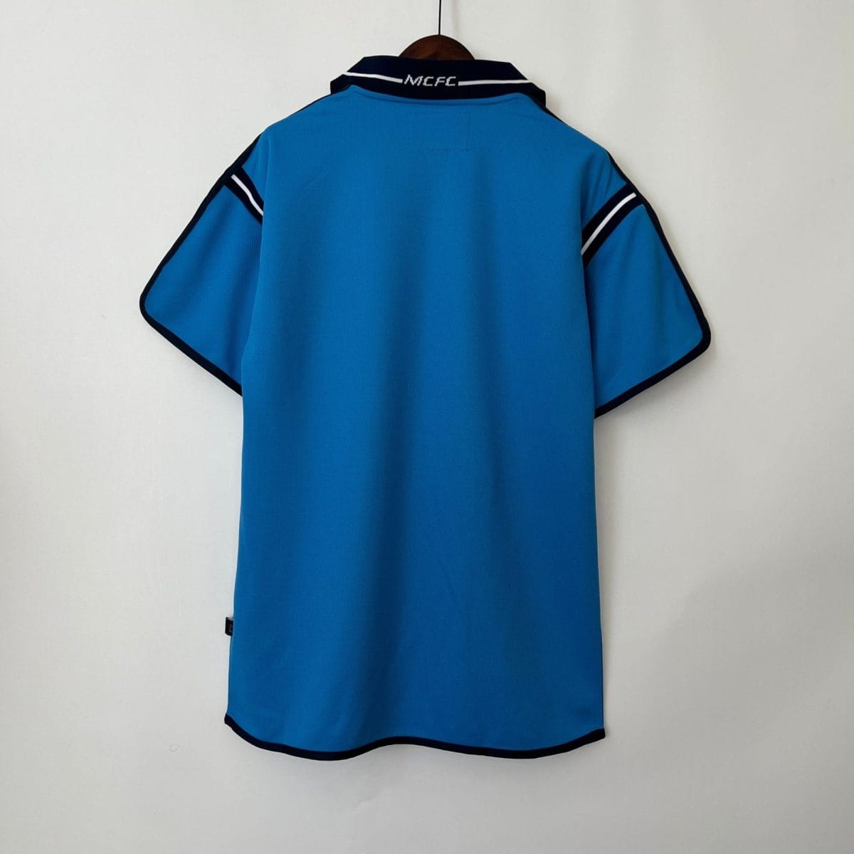 Back view of a bright blue polo jersey on a wooden hanger, collar reads MCFC with navy trim at edges.