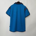 Back view of a bright blue polo jersey on a wooden hanger, collar reads MCFC with navy trim at edges.