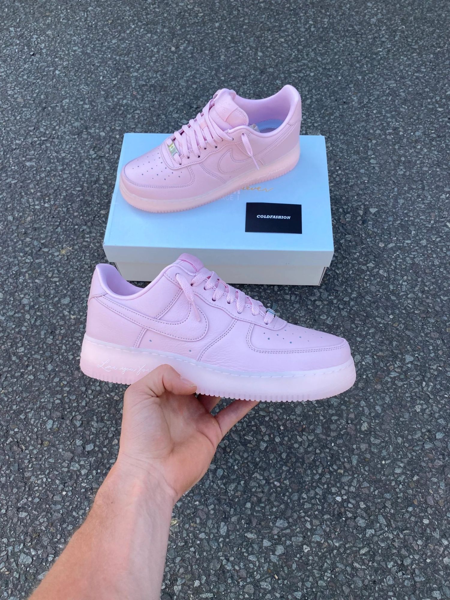 87F6654C-44D0-47CB-A1A6-532D40B39CD0 | UAE SPORT STORE Pink low-top sneakers resting on a light blue box on a rough asphalt surface, showing Nike swoosh and laces clearly.