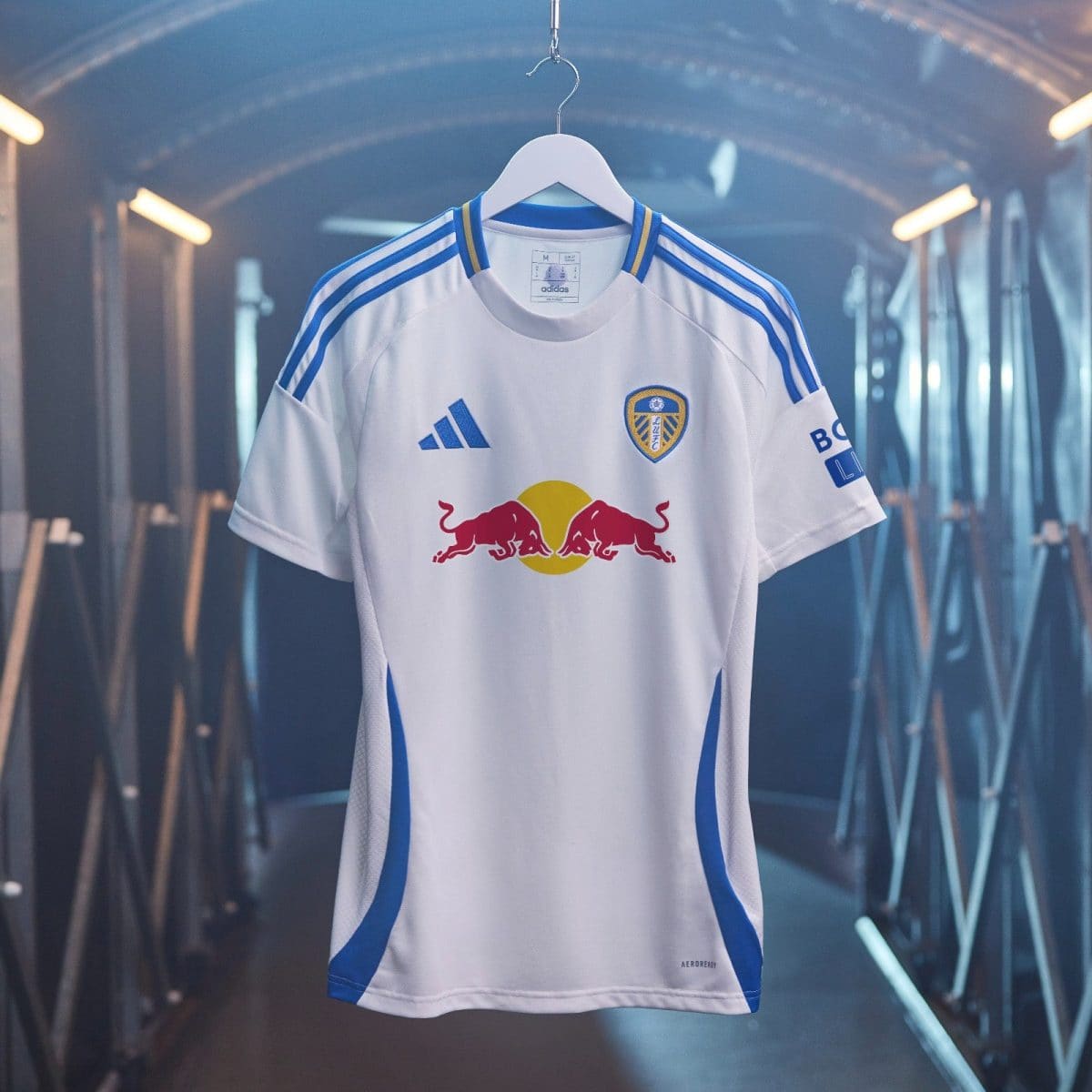 White Leeds United football jersey on a hanger in a dimly lit tunnel, featuring blue Adidas stripes and the Red Bull logo across the front.