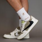 Beige and white sneakers with a black accent, worn on a person’s feet, paired with white crew socks and a gray background.