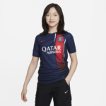 880b5f33_93a4_4f43_86a7_7408127c8445 | UAE SPORT STORE Young woman modeling a Paris Saint-Germain soccer jersey in navy blue with a red vertical stripe and Qatar Airways sponsor, standing with one hand in her pocket against a light background.