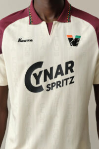 Cream-white sports jersey with burgundy shoulders and a V-notch collar; features Nocta logo on the left chest and a stylized team crest on the right, plus a large 'Cynar Spritz' sponsor graphic across the front.