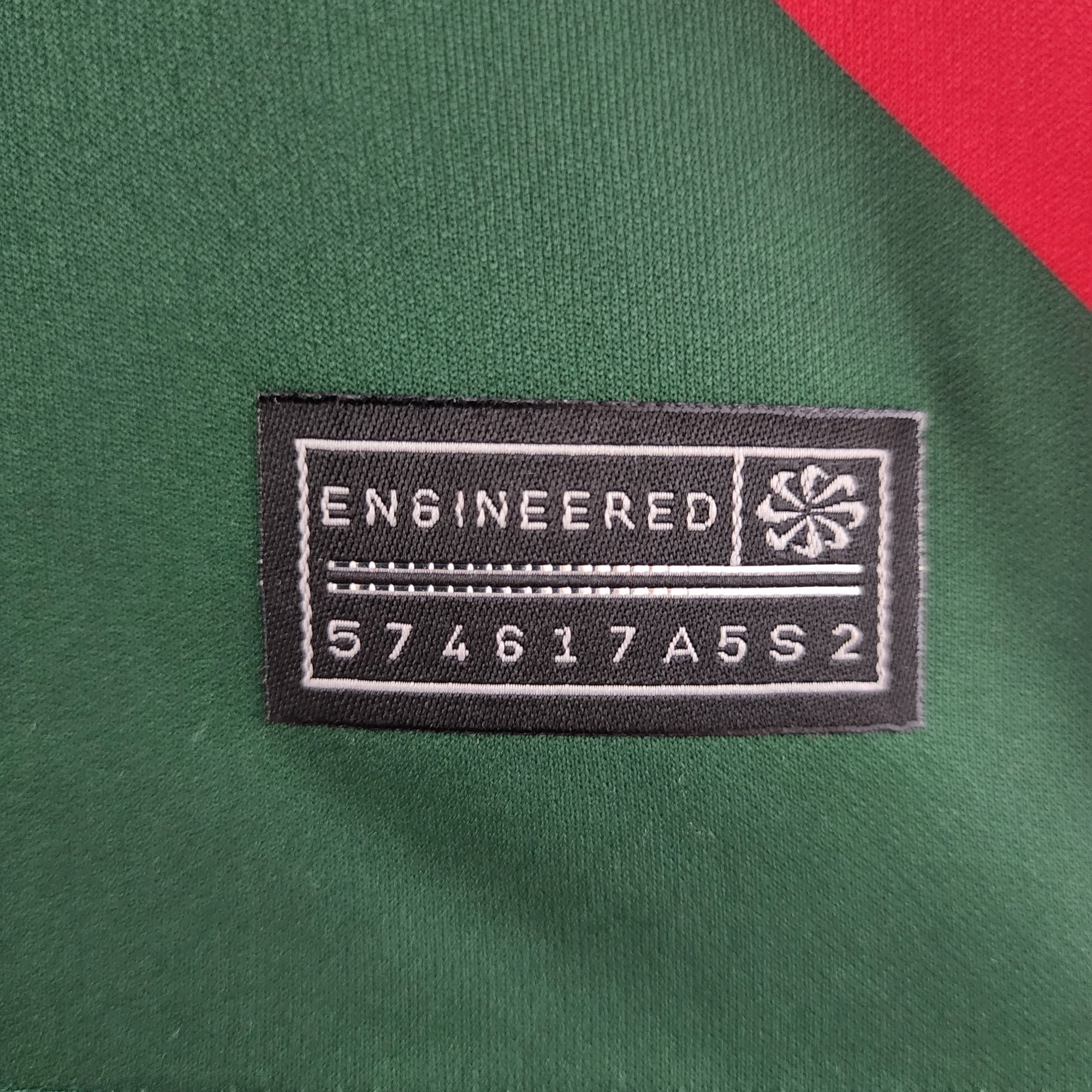 890e12ab_971adba7-d5a6-4c32-8b76-0256bf9f31af | UAE SPORT STORE Close-up of green fabric with a black rectangular label reading 'ENGINEERED' in white, bordered by white stitching and a red diagonal accent in the corner.
