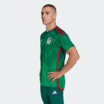 Male model wearing green Adidas soccer jersey with red piping and team crest, standing in profile with hands on hips.