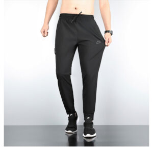 Black Nike men's joggers with drawstring waist and tapered legs, shown on a model with sneakers and a wristwatch in a studio setting.