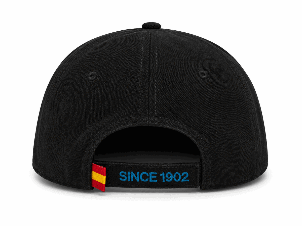Back view of a black baseball cap with an adjustable strap, featuring blue 'SINCE 1902' embroidery and a small red-yellow flag tab