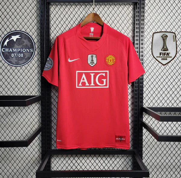 Red Manchester United home jersey on a hanger, featuring the AIG sponsor, Nike swoosh, and club badges with Champions patches nearby.