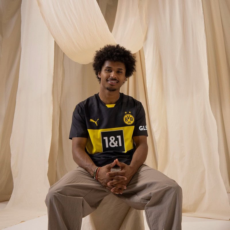 8CfH3sM1b9aJn3U | UAE SPORT STORE Portrait of a man with curly hair, sitting cross-legged in a Borussia Dortmund jersey against beige drapery.
