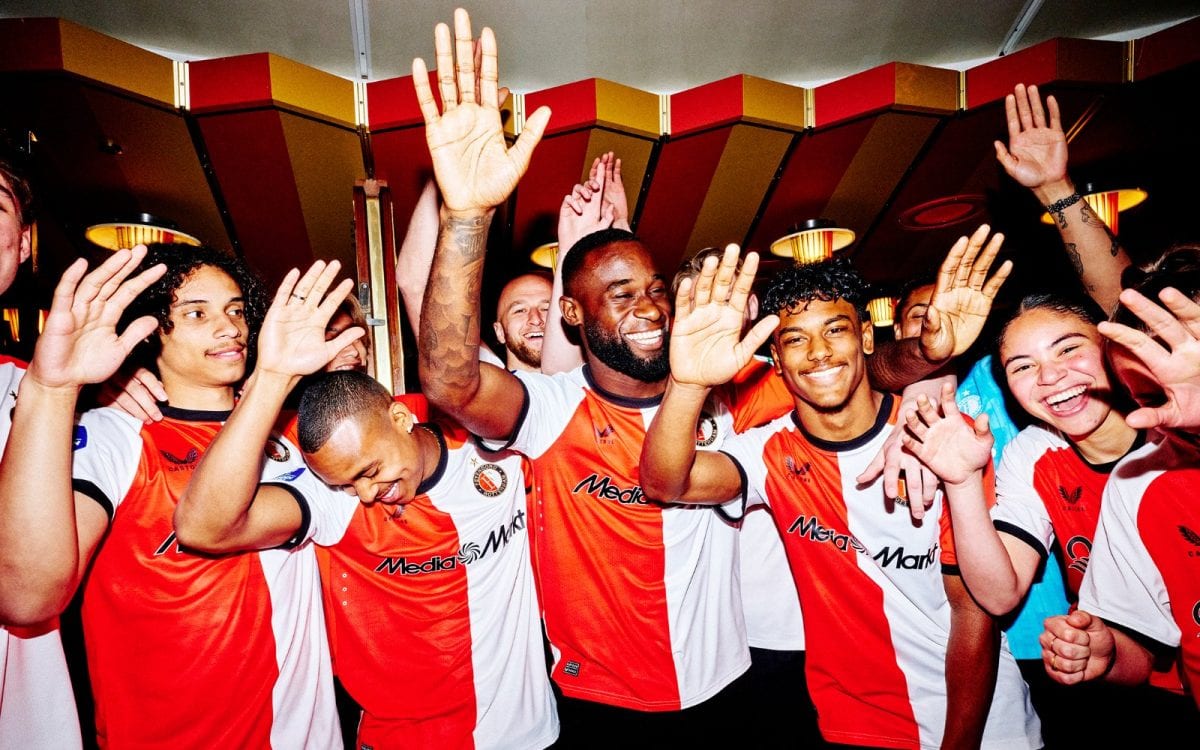 A diverse group of soccer players in red-and-white jerseys cheer with hands raised in celebration indoors.