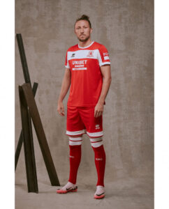 Male soccer player in a red kit with white trim and Unibet sponsor, posing indoors beside a wooden frame.