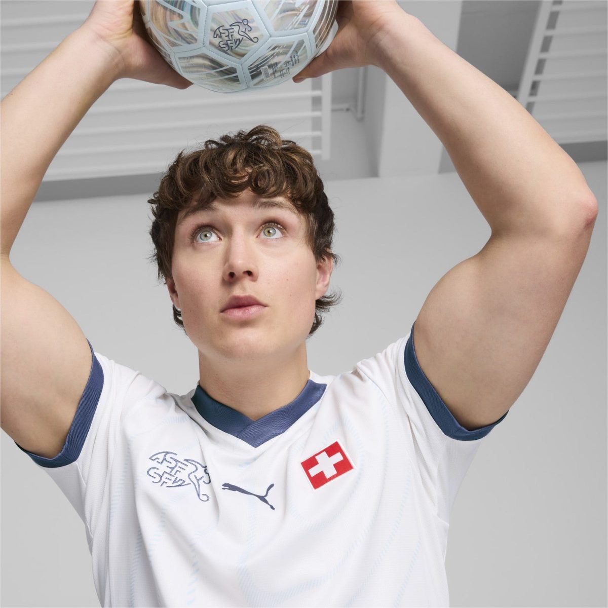 Young athlete in a white jersey with Swiss flag, holding a soccer ball overhead and looking up, indoors.